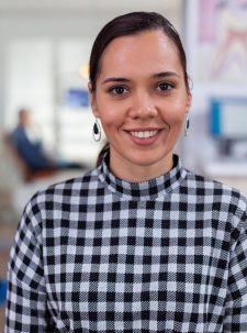 Portrait of smiling woman patient looking on camera sitting on chair in waiting room of stomatological clinic. Stomatologist assistant typing on pc in dental office while doctor working in background.